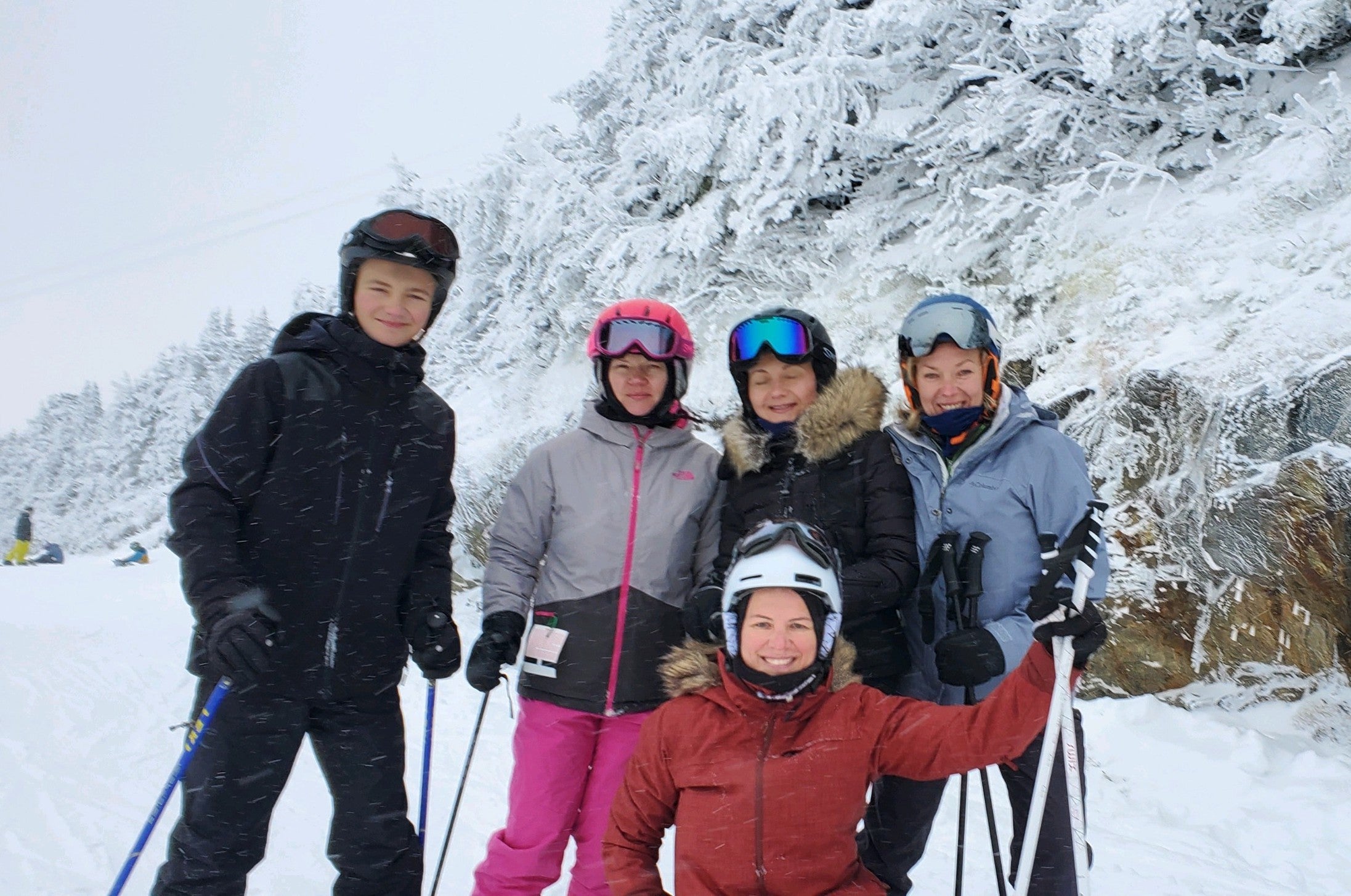 Family ski vacation, 5 people wearing winter ski clothing and holding ski poles, posing for a group photo with snow covered trees in the background.