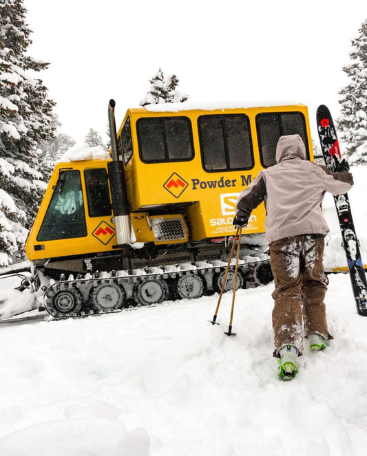  Powder Mountain activity. Person holding skis walking toward big snow cat vehicle that looks like a tank. Trees covered in snow in background.