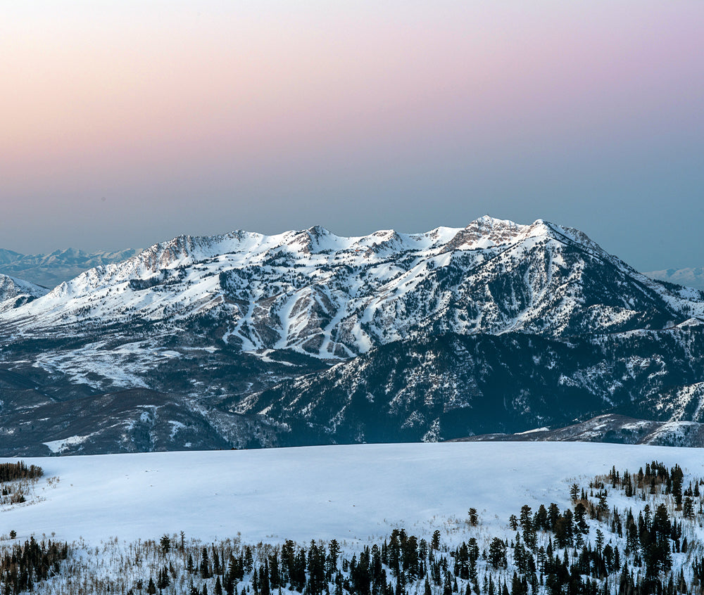 Huge mountain and field covered in snow at the edge of a pine forest. Pink and hazy sky during sunset at Snowbasin.